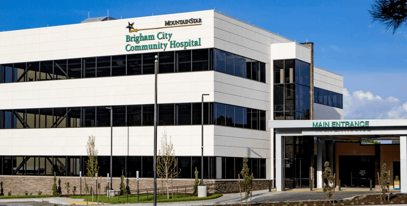 Exterior view of Brigham City Community Hospital, part of the MountainStar network, showing a modern white building with large windows and a sign for the main entrance.