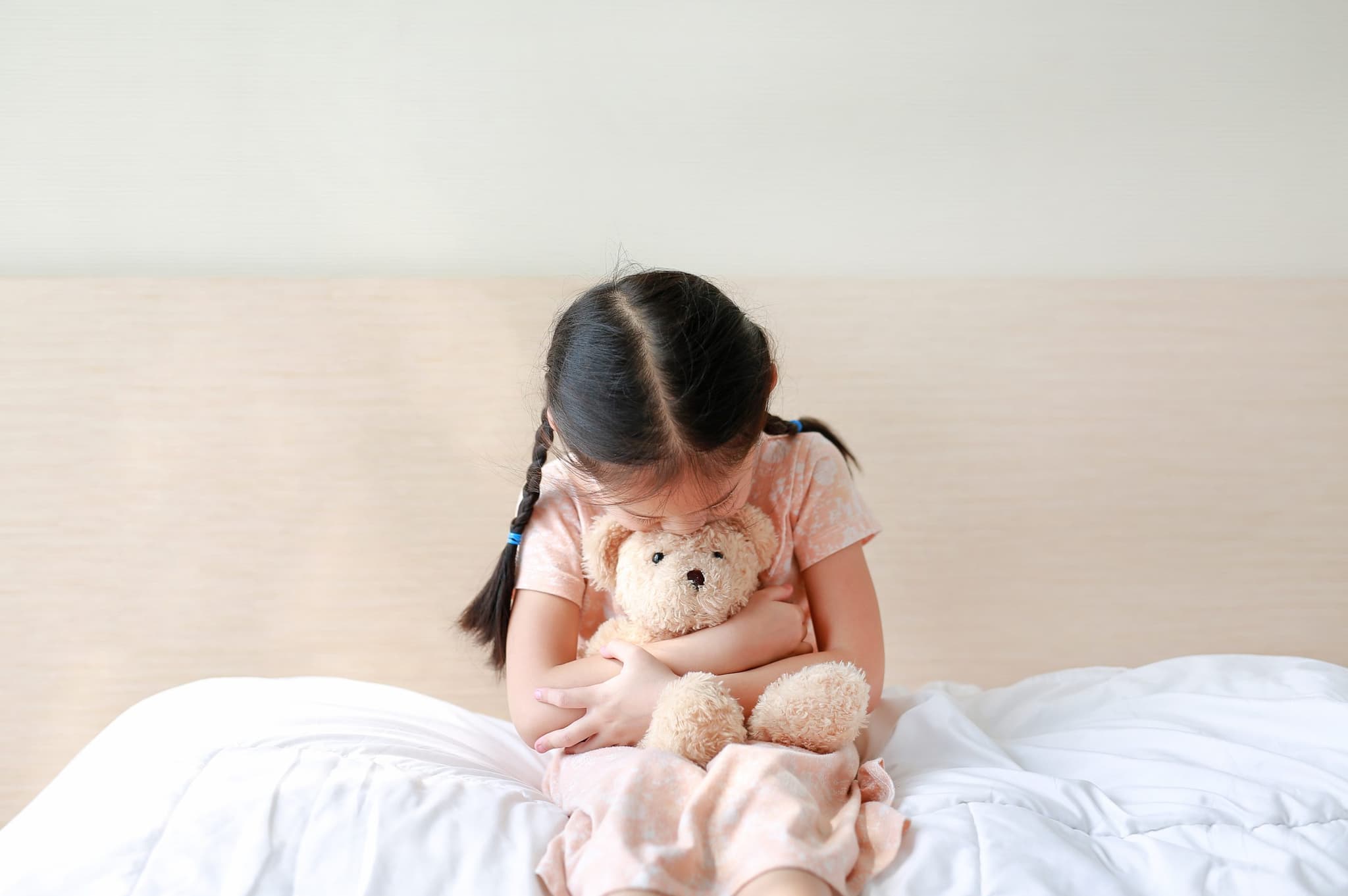 Picture of Peaceful little girl embracing teddy bear while sitting on the bed at home.