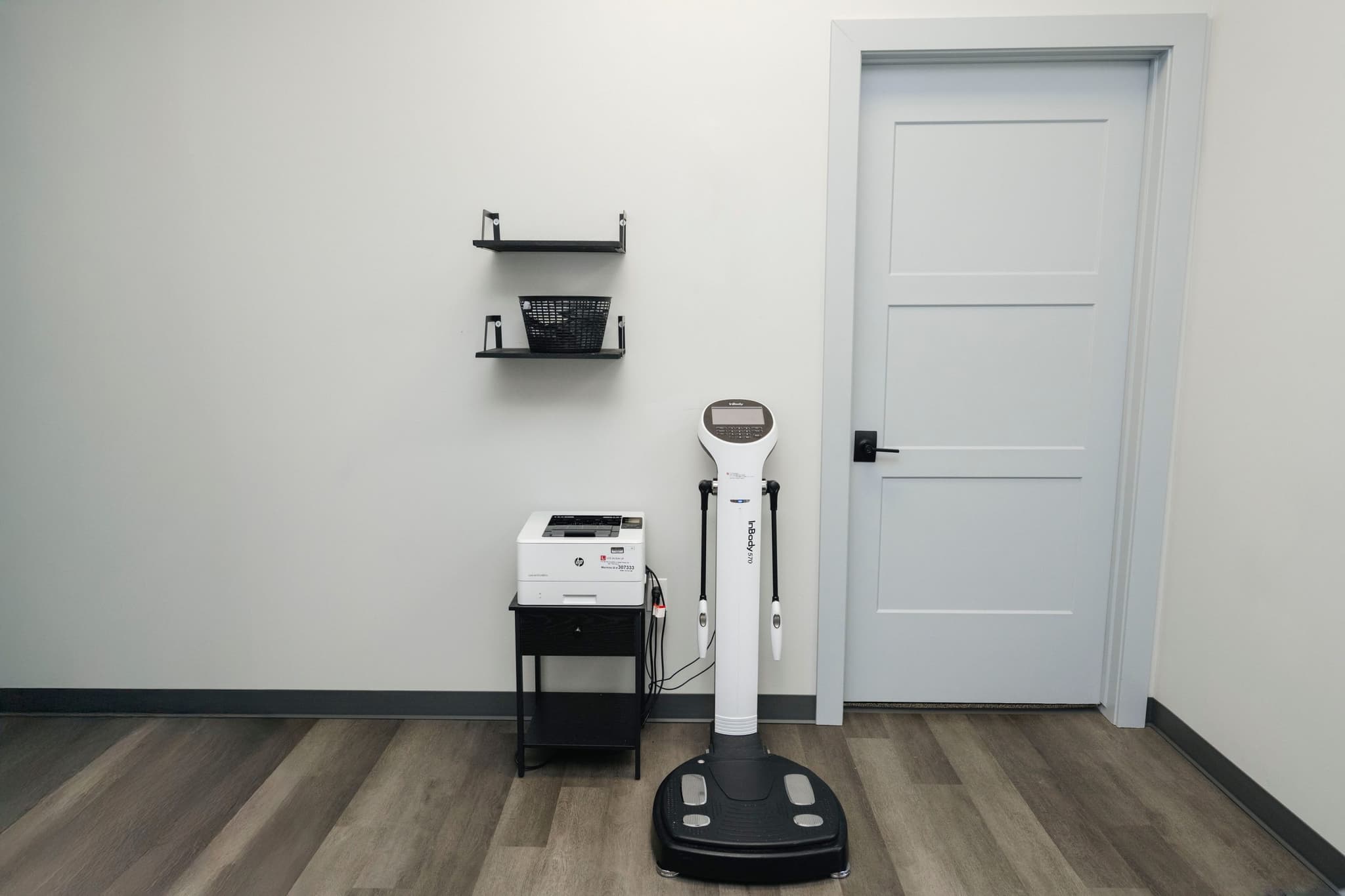 Image of A clean clinical room with an InBody scan machine used for body composition analysis, positioned beside a small shelving unit and a closed door.