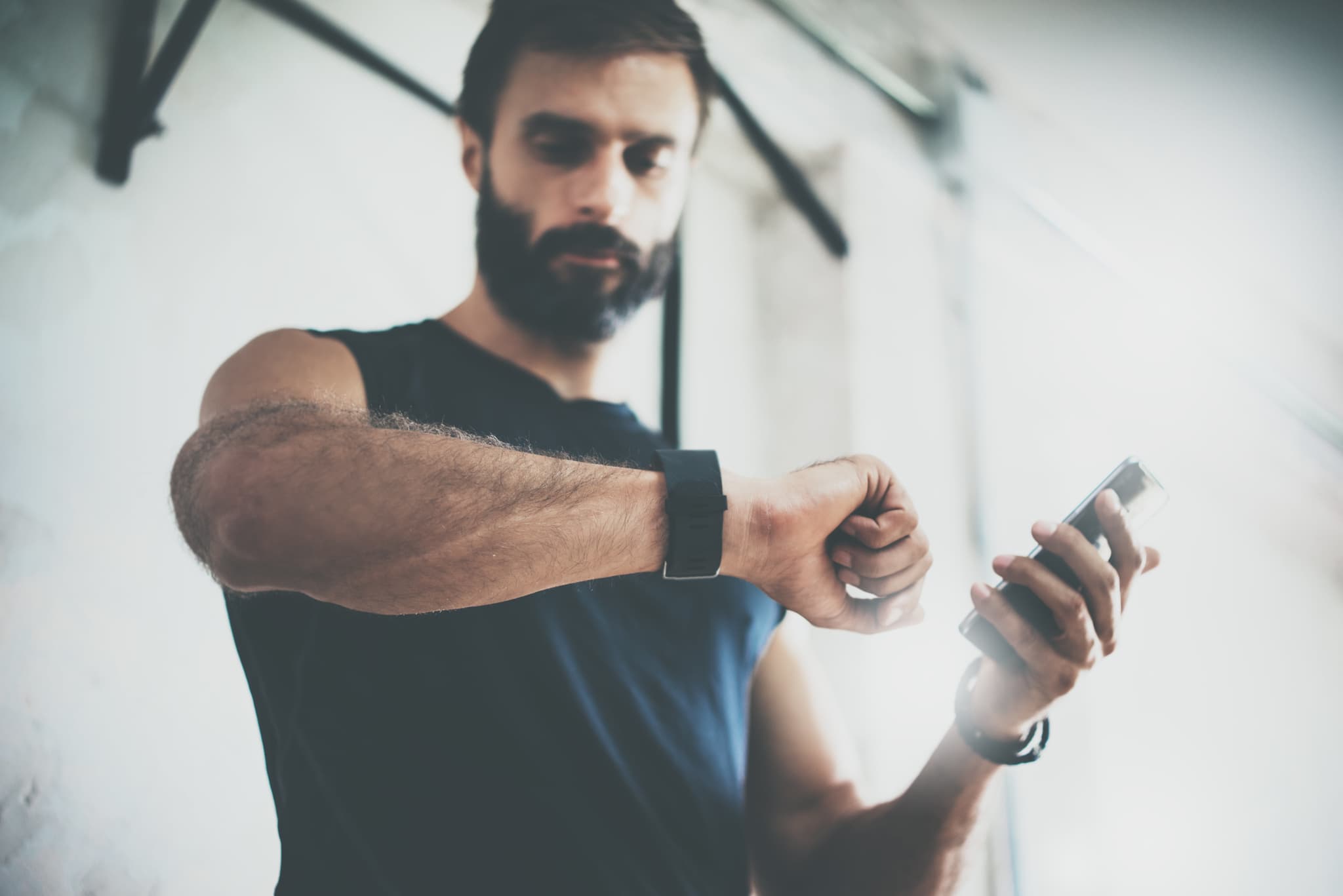 Photo Bearded Sportive Man After Workout Session Checks Fitness Results Smartphone. Adult Guy Wearing Sport Tracker Wristband Arm. Training hard inside gym. Horizontal bar background. Blurred