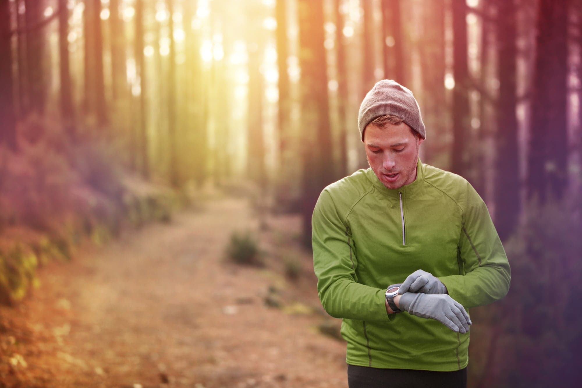 Picture of Trail running runner looking at heart rate monitor watch running in forest wearing warm jacket sportswear, hat and gloves. Male jogger running training in woods.