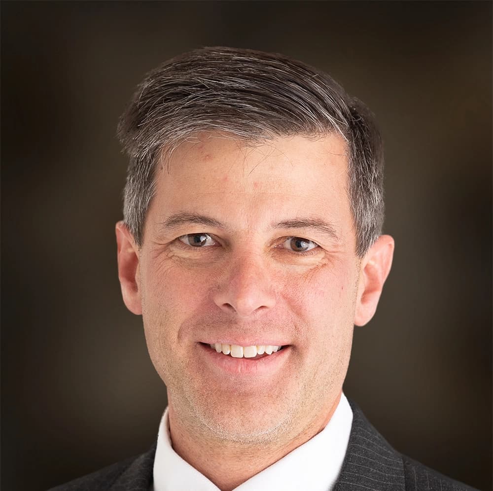 Portrait of Edward Prince, MD, a man with short gray hair, wearing a suit and smiling against a brown background.