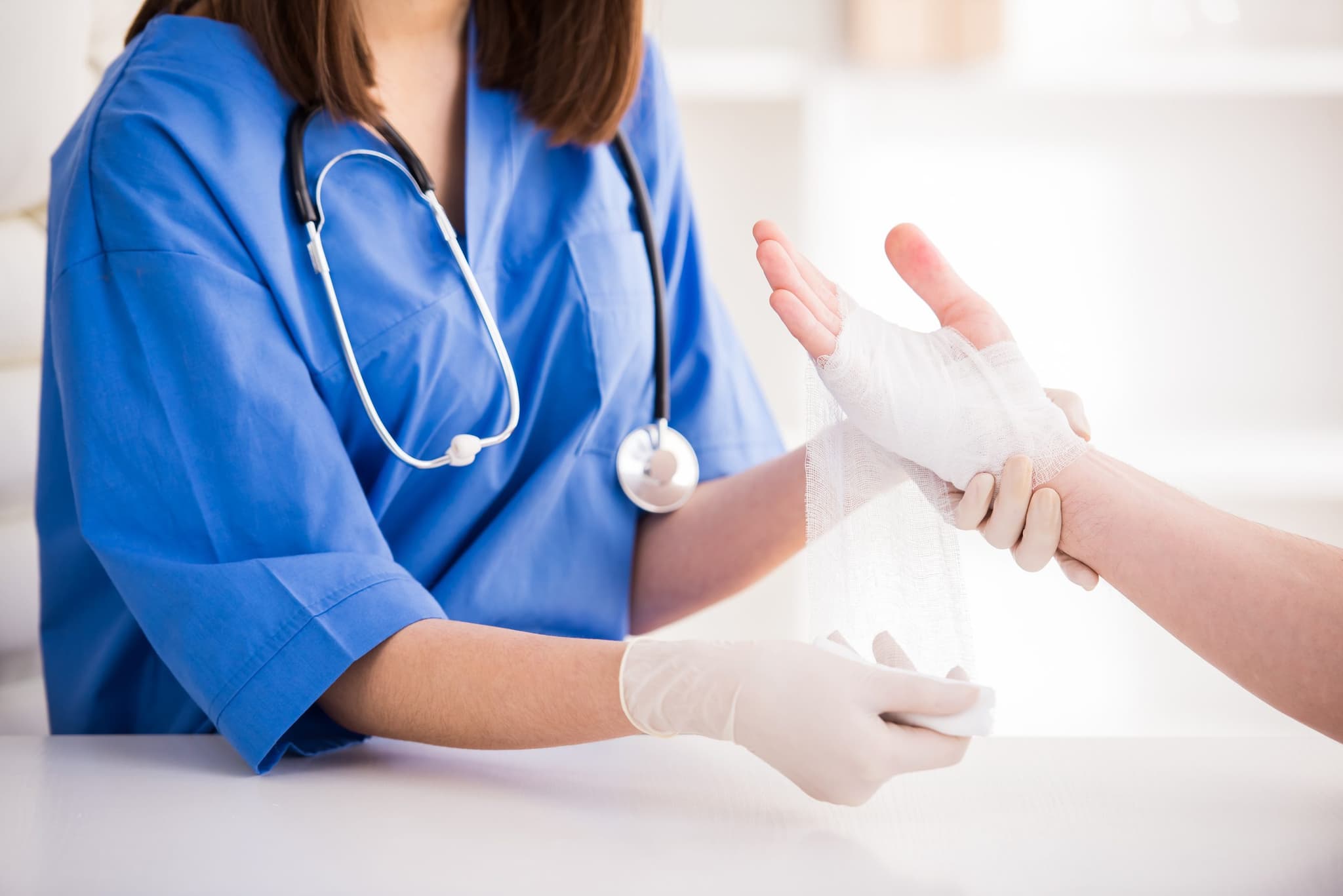 Picture of A healthcare professional in blue scrubs and latex gloves bandaging a patient's injured hand with gauze, providing medical care in a clinical setting