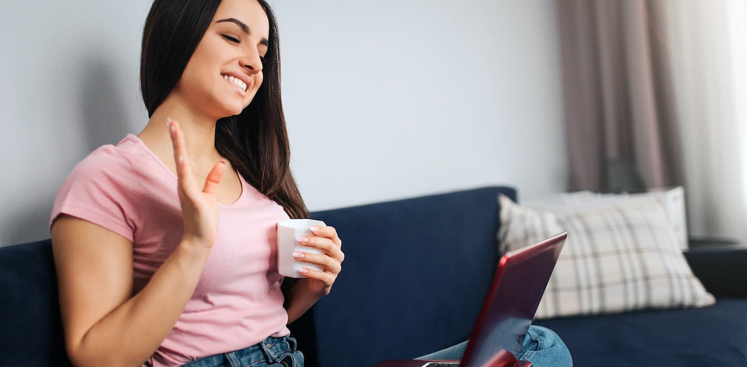 Image of Cheerful young woman sit on sofa and wave with hand. She smiles and look on laptop screen. Model hold it on nap. She has white cup of drink.