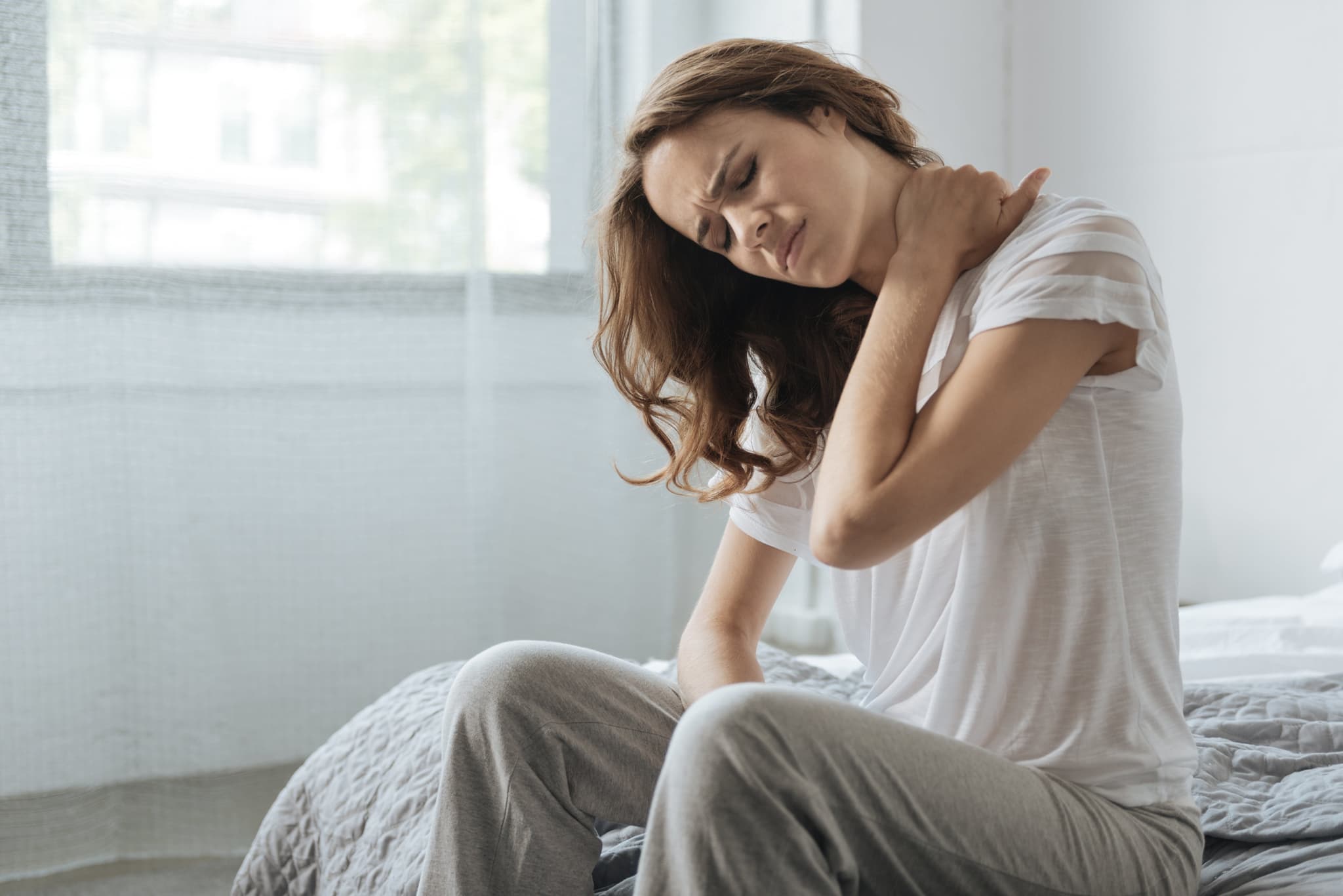 Picture of Neck inflammation. Moody unhappy young woman sitting on the bed and holding her neck while having an inflammation there