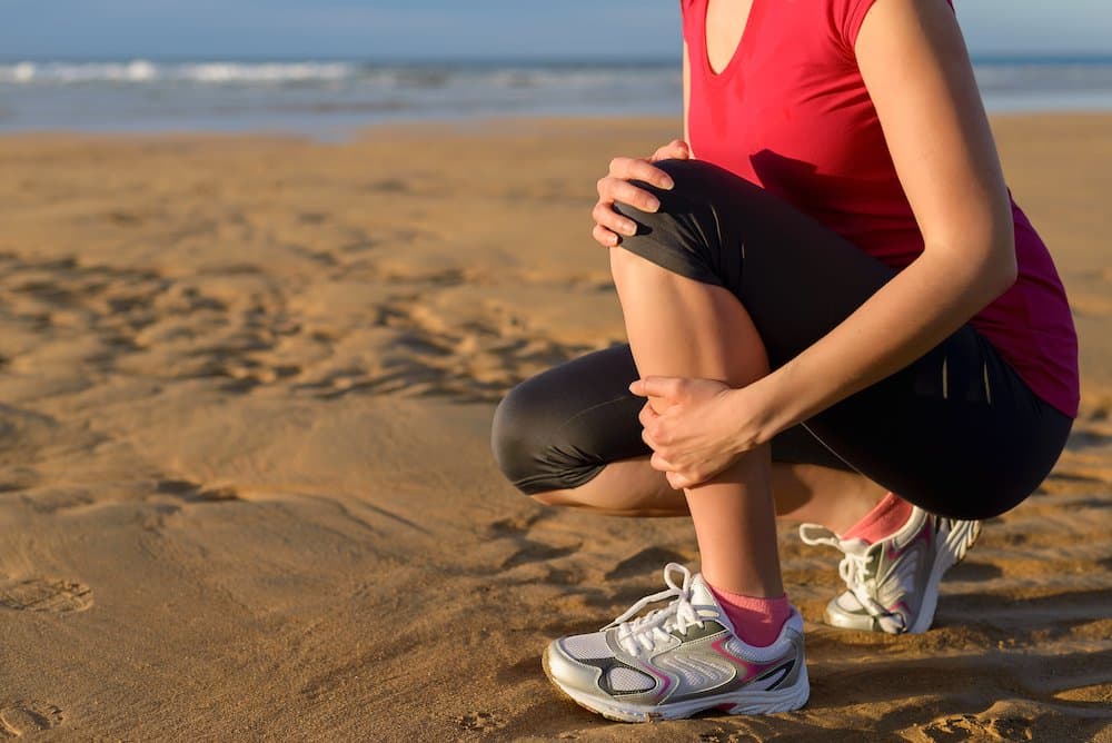 Picture of Female runner clutching her shin because of a running injury and inflammation. Tibial periostitis hurt while jogging on beach.