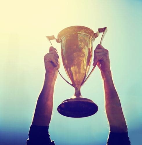 Picture of a man holding up a gold trophy cup as a winner in a competition toned with a retro vintage instagram filter effect app or action (backlit with the sun)