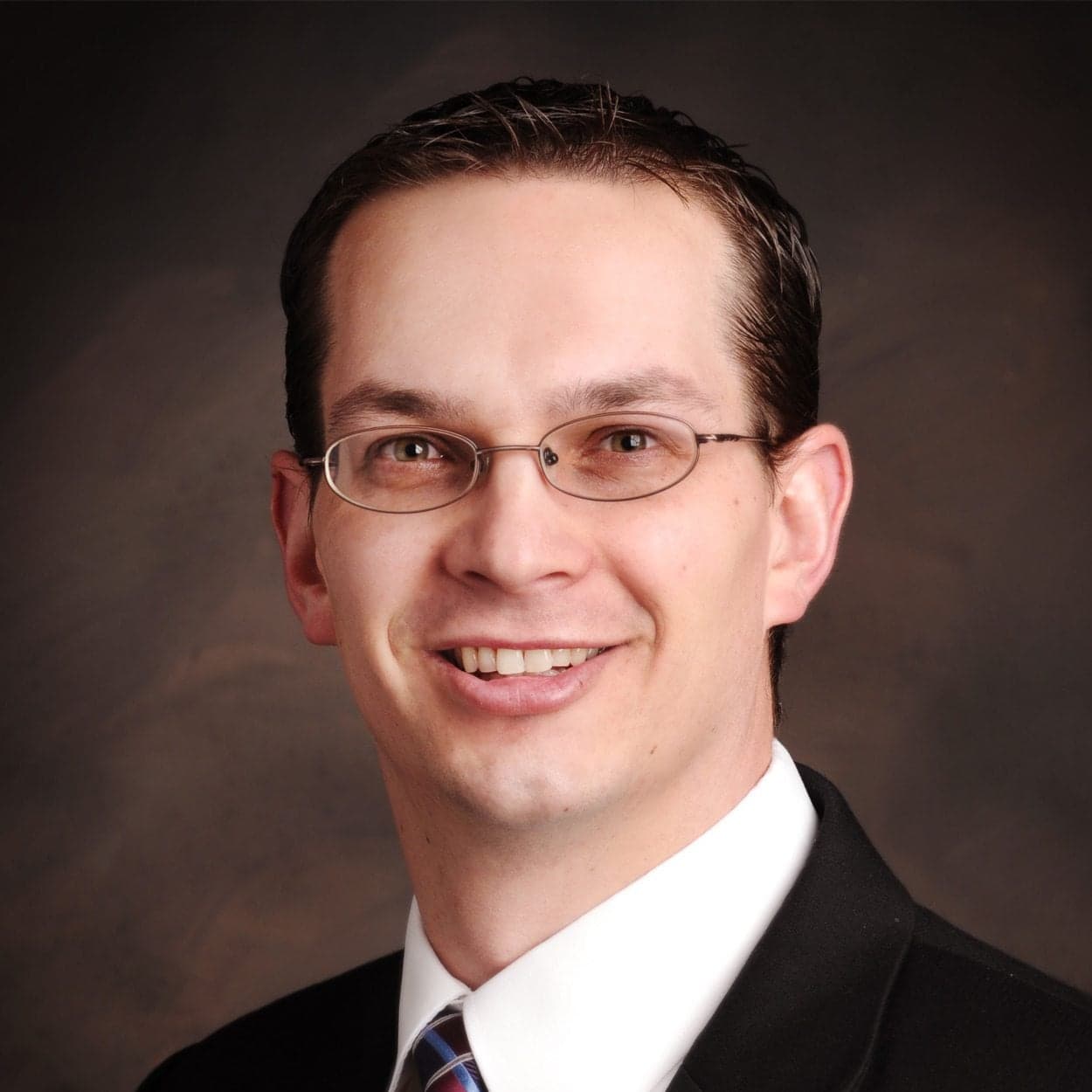 Portrait of Garry Miller, DO, smiling with short dark hair and glasses, wearing a suit and tie, against a dark brown background.