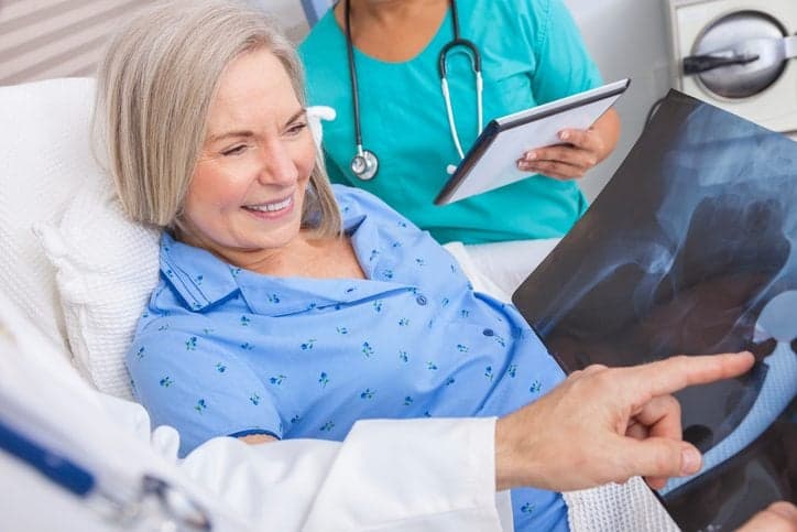 Happy senior woman patient recovering in hospital bed with male doctor and female nurse looking at hip replacement x-ray