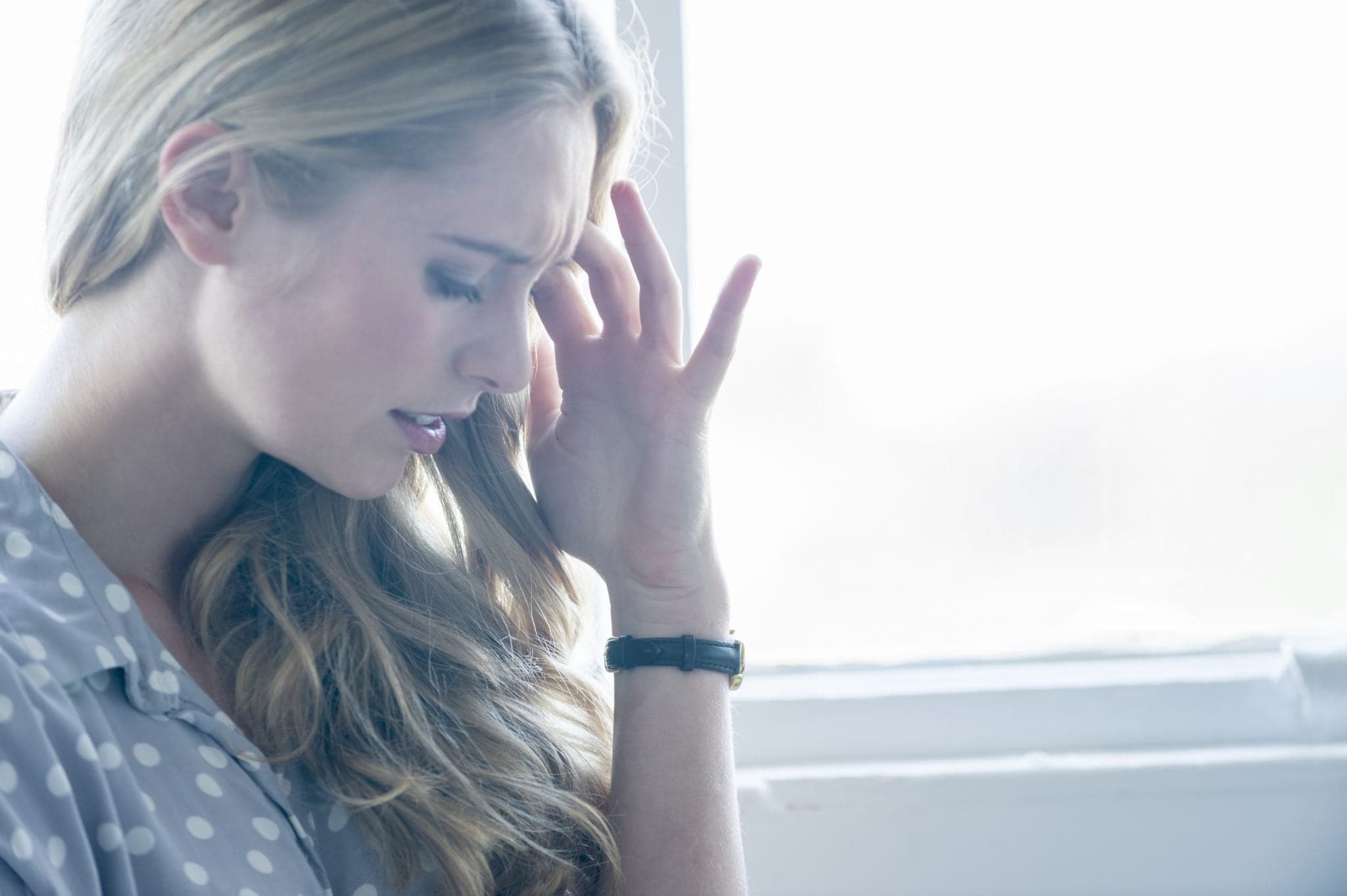 Picture of Woman looking upset or in pain. Attractive woman leaning against a wall with her hand on her forehead. She could be in pain, upset, angry, depressed or stressed. She is frowning and standing in front of a window. She is casually dressed.