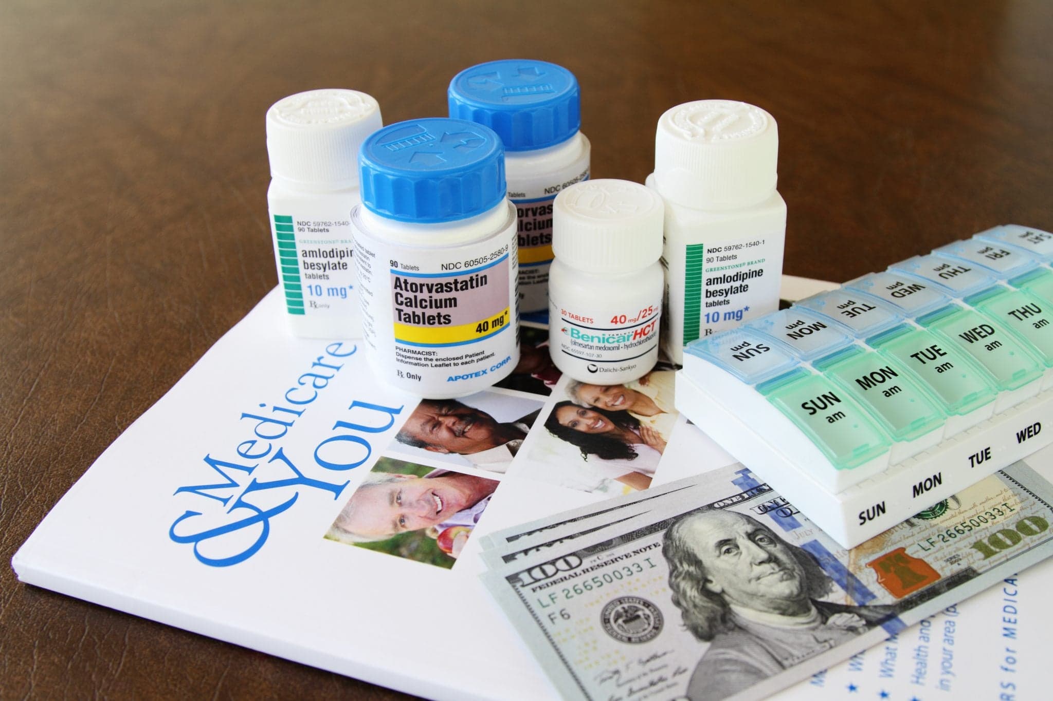 Image of Prescription bottles containing various prescription medicnes for high blood pressure and cholesterol are grouped on the publication. A compartmentalized daily medicine dispenser is also on top of the booklet. Several one hundred dollar bills are partially visible as well.