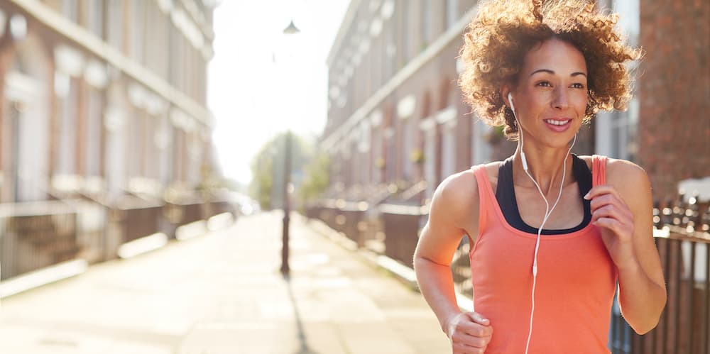 Picture of a young woman running in a street on her morning or evening run . She is backlit by a rising low sun , and is pictured staring ahead with determination , before heading off on her morning jog.She is wearing a vest top , trainers and leggings , and earphones. The sun is rising at dawn behind her or setting at sunset . Lens flare streaks across the frame .