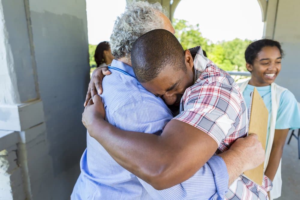 Emotional African American father embracing the caucasian senior man in a light blue shirt. His daughter is standing smiling behind them, she is wearing a light blue shirt with a vest on top of it.