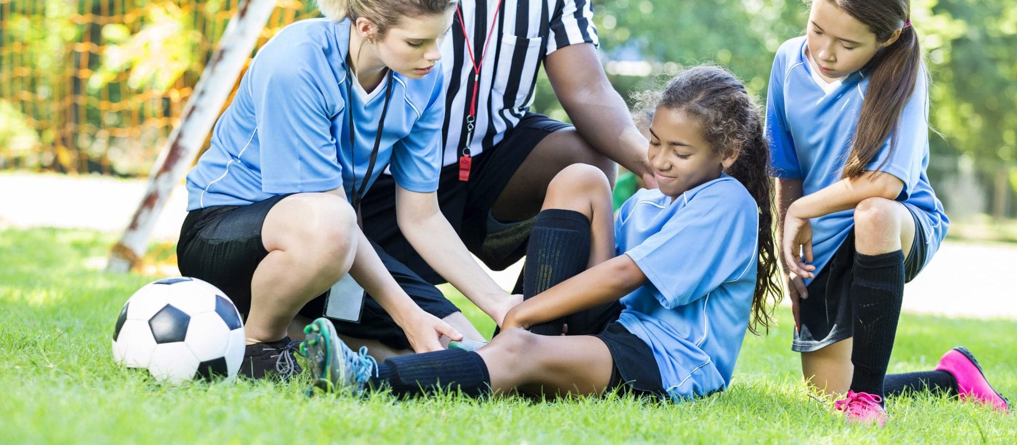 Picture of Young girl soccer plyer with an injury, she is hurt on the sidelines. The coach and referee are checking her injuries, her concerned friend and teammate is kneeling on a knee next to her friend. The girls are wearing matching light blue soccer uniforms with black shorts. Their referee is wearing a white and black striped referee uniform.