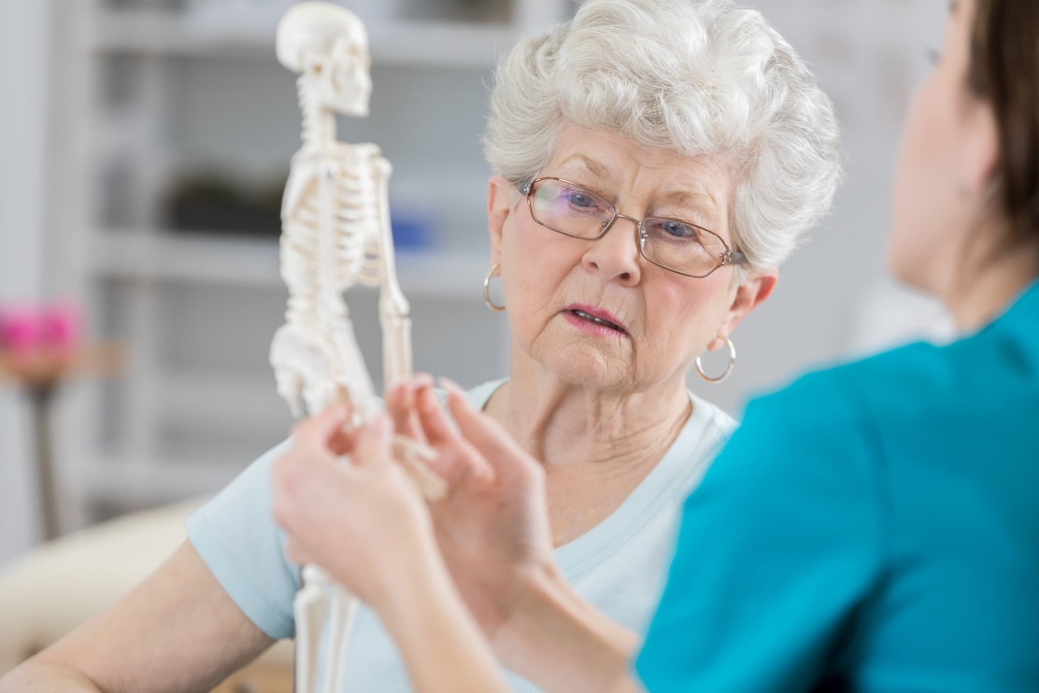 Senior woman listens seriously as her physical therapist sits with her and holds a model of the human skeletal system. The therapist is discussing the affects of osteoporosis in posture and the reason for resulting pain.