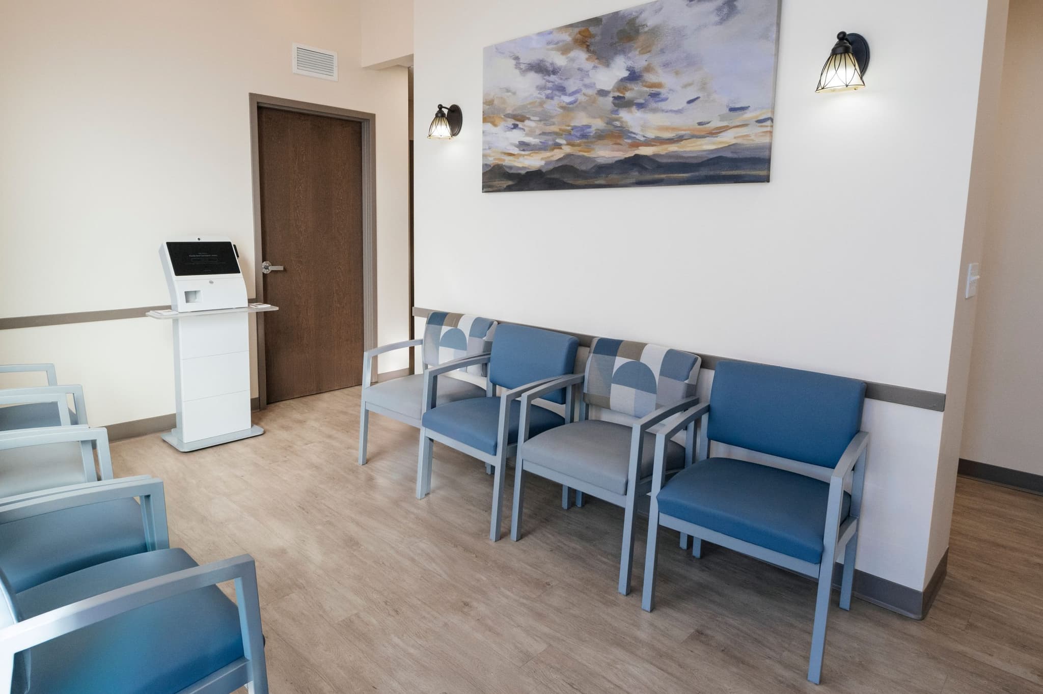 Image of Bright waiting area with blue cushioned chairs, light wood flooring, and a landscape painting on the wall.