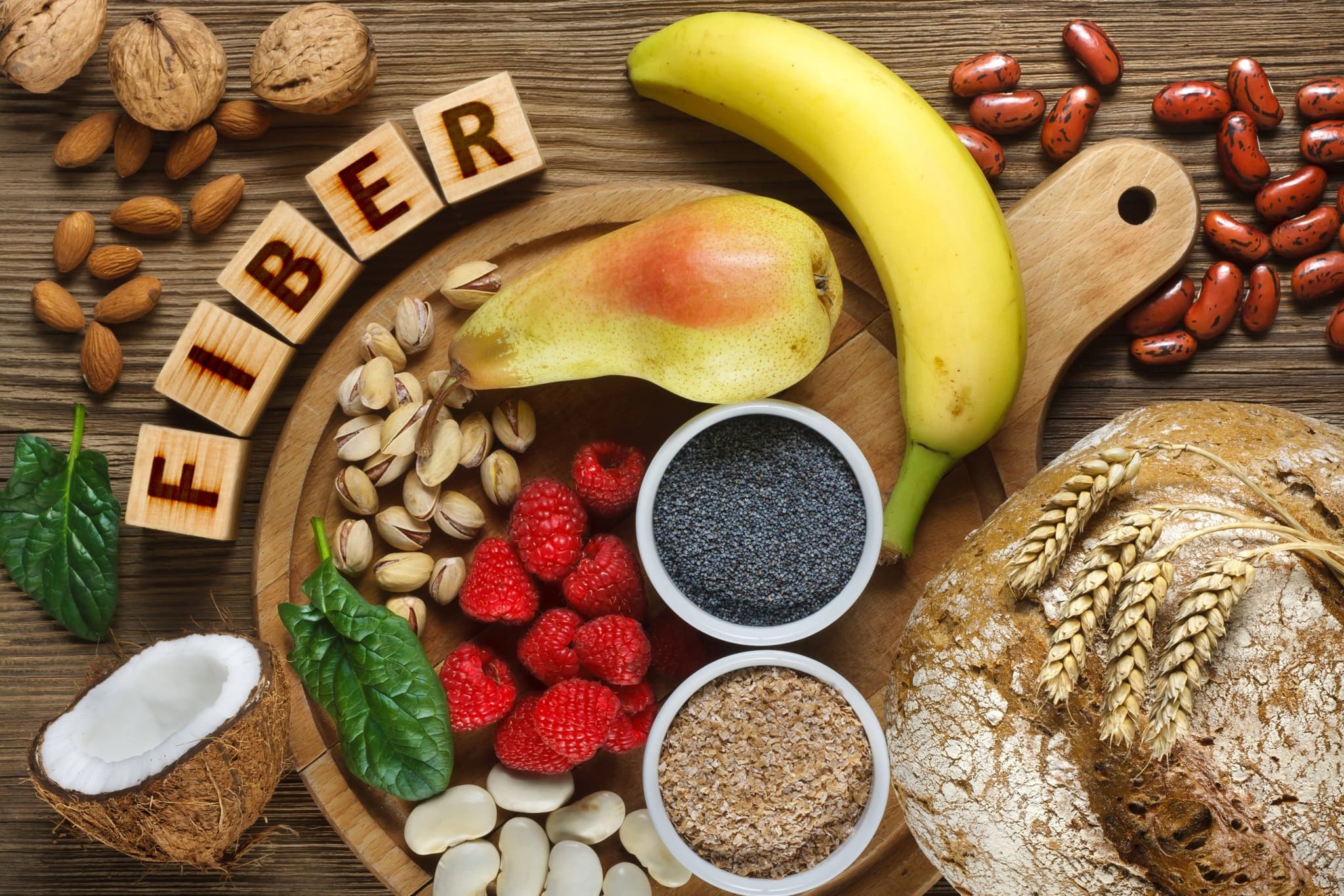 Picture of Foods rich in fiber as rye bread, wheat bran, white beans, red beans, spinach, almonds, poppy seed, pears, bananas, coconut, raspberries, pistachios, walnuts. Wooden table as background