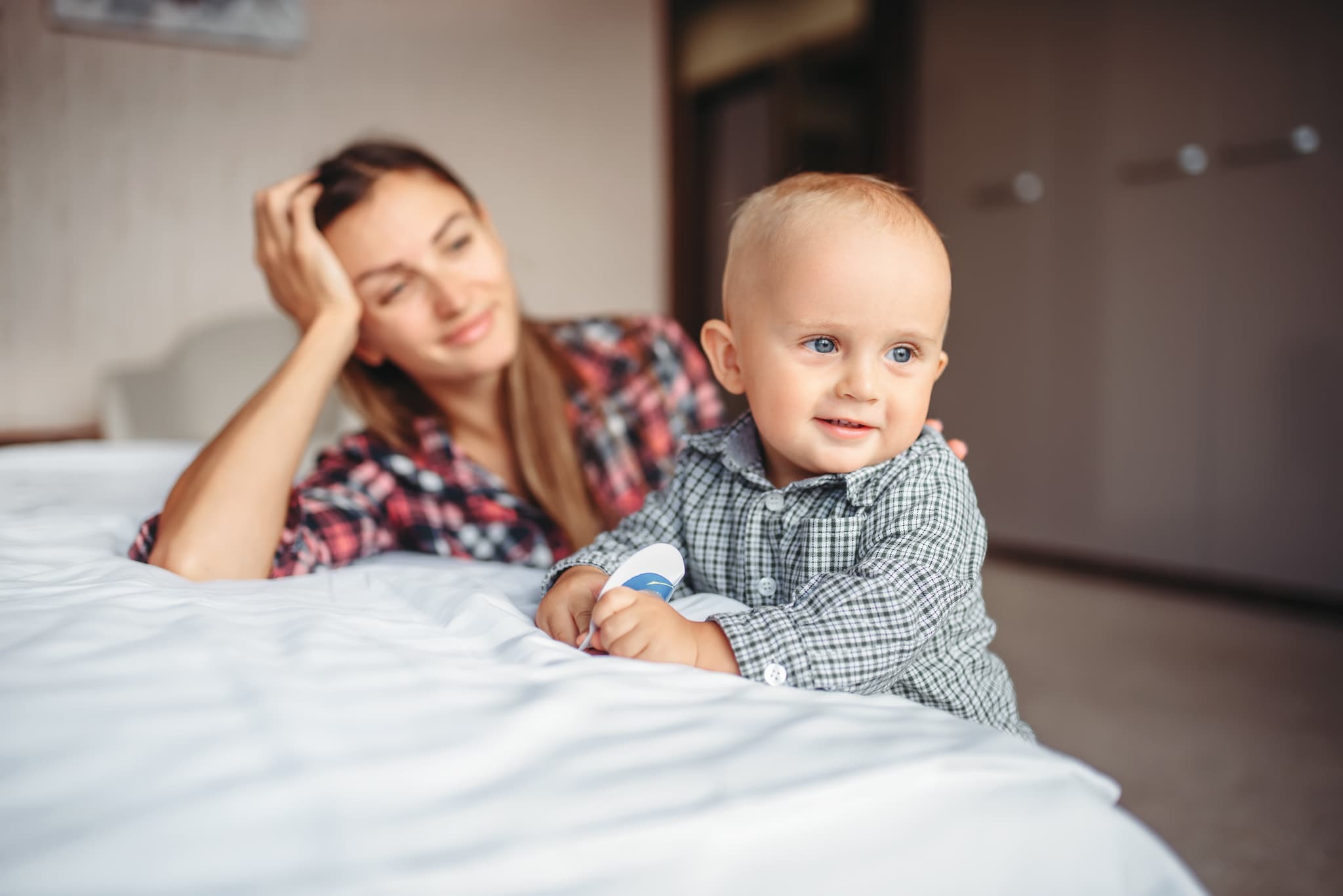 Tired mother looks on her little child playing on the bed, motherhood problems. Depressed mom and son together at home, parenthood trouble